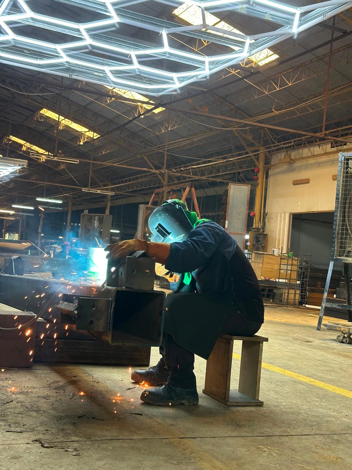 Prior plant work — welder on a large steel beam under shop lights, San Simon (not a Calma.Basic product)